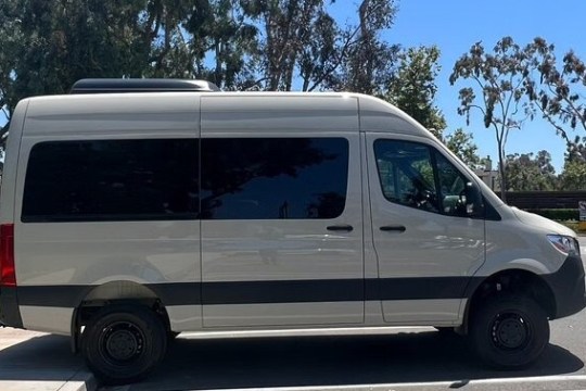 White cargo van parked on street beside a park with trees and a clear sky.