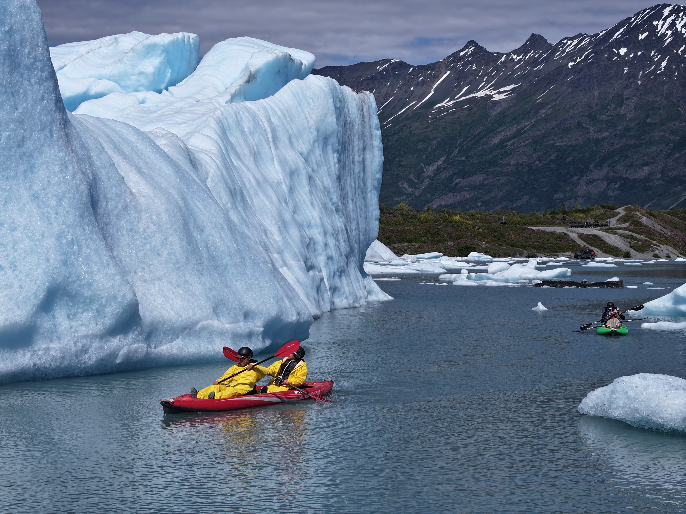 Kayakers in bright gear paddle near large icebergs with mountains in the background.