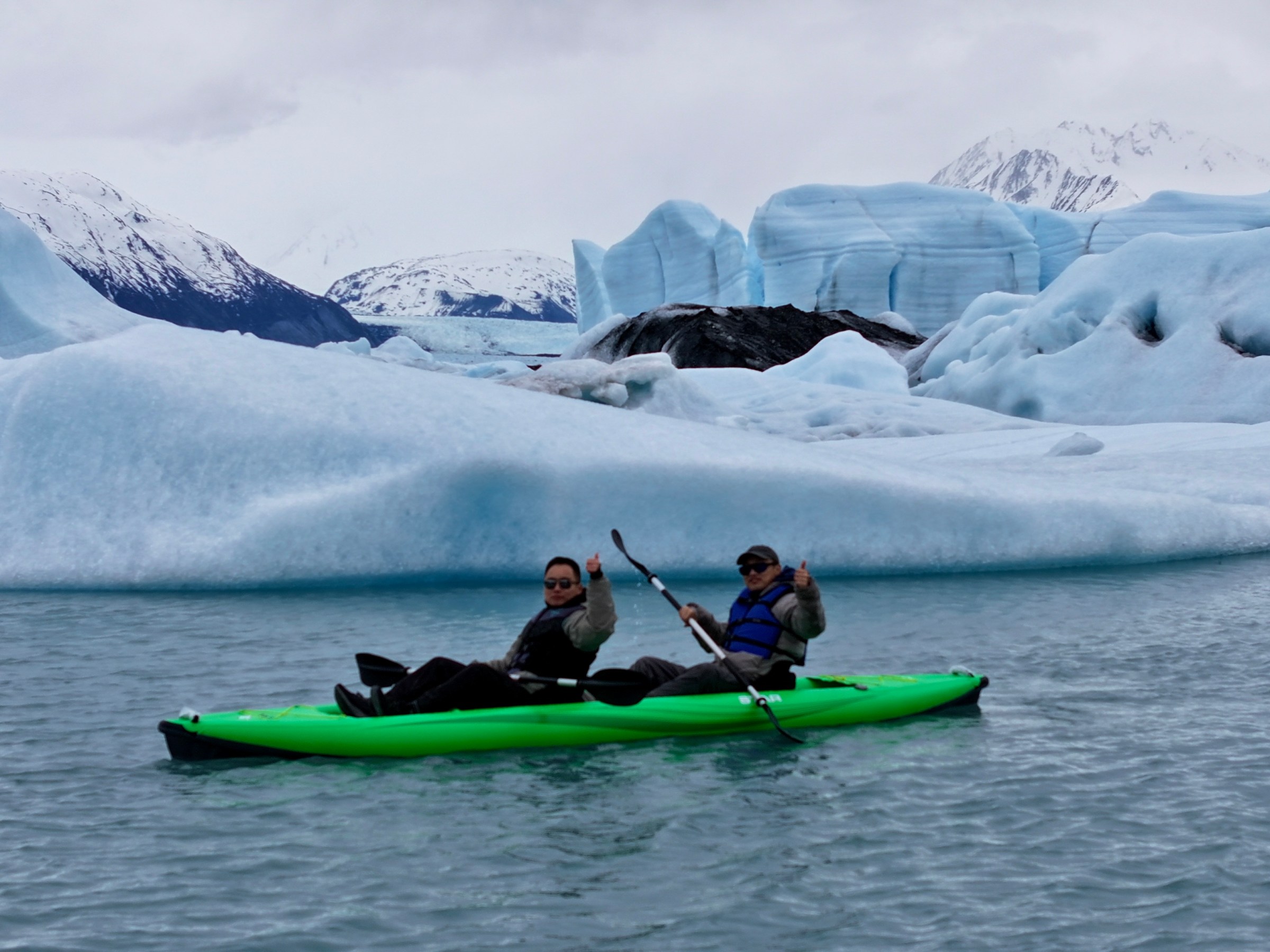 Two people kayaking in a lake surrounded by icebergs and snow-covered mountains.